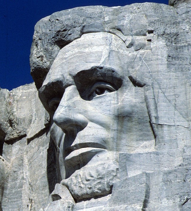 Closeup view of Abraham Lincoln's image carved onto the gray granite on Mount Rushmore.  He is looking to the left with a pleasant or perhaps thoughtful expression.  Lighter colored veins of granite and cracks are visible crossing his forehead and nose.