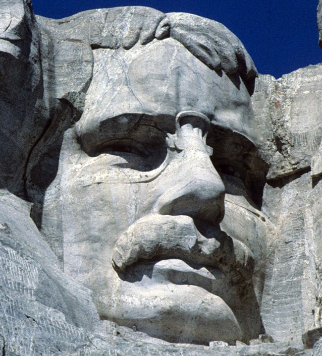 View of Theodore Roosevelt's image carved onto the gray granite on Mount Rushmore.  He is looking slightly to the right and straight ahead and is depicted with a stern expression.