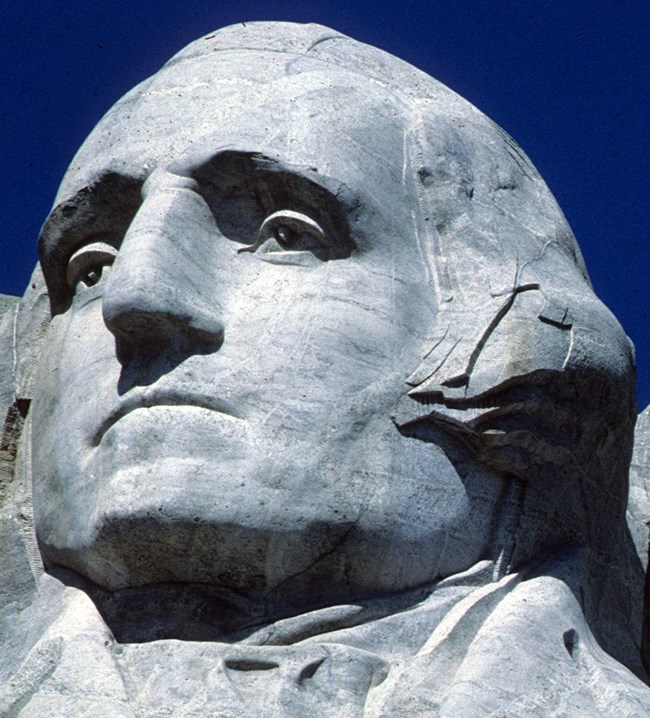 Closeup view of George Washington's image sculpted onto the gray granite of Mount Rushmore.  He is looking slightly to the left and depicted with a stern expression.