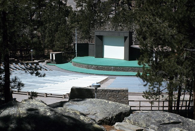 View of the original amphitheater built in 1959 from the rear of the seating area.  A small screen is surrounded by brown wood creating a stage.  The stage is green.  Numerous gray colored benches form an arc around the stage.