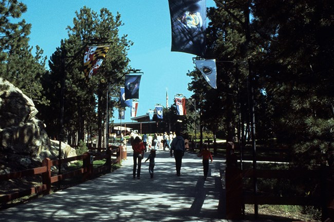 Visitors walking from the parking lot to the Visitor Center along the Avenue of Flags as it looked in 1976.  A red fence and ponderosa pine trees line the path and state flags hang vertically about ten feet above the walkway.