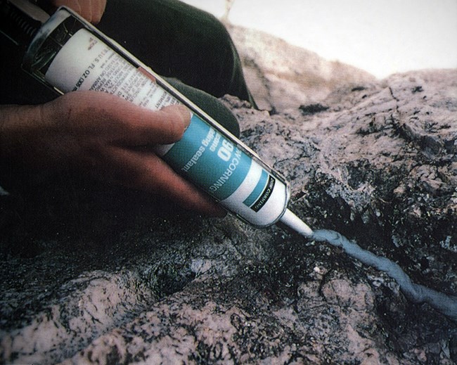 A worker in the upper left of the image applies silicone caulking with a caulking gun to a crack in the lower right on Mount Rushmore.