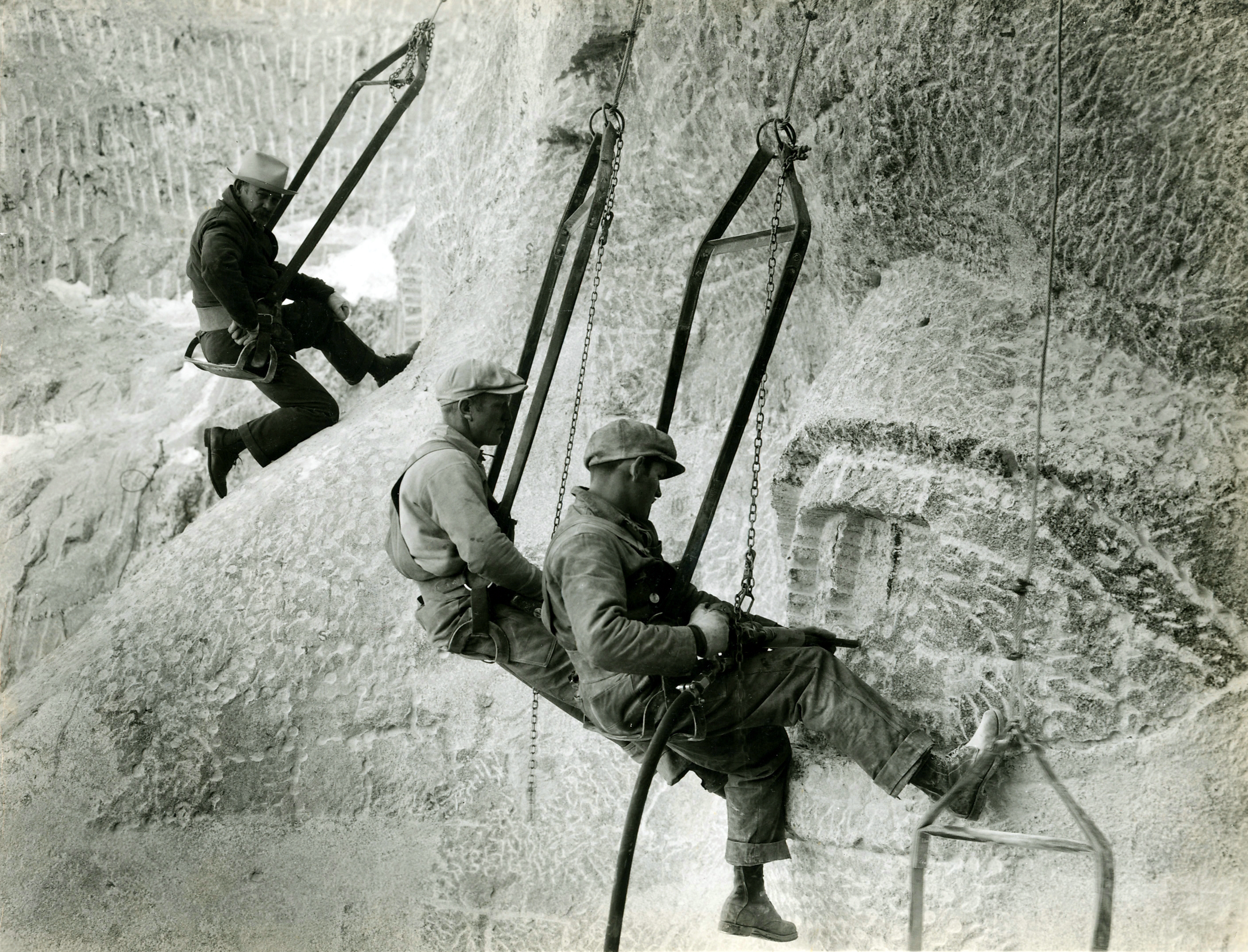 Gutzon Borglum on a sculpted nose in the upper left of the image observes two workers in the bottom center carving an eye on Mount Rushmore.  All 3 are in bosun's chairs.  The two workers are using pneumatic drills.