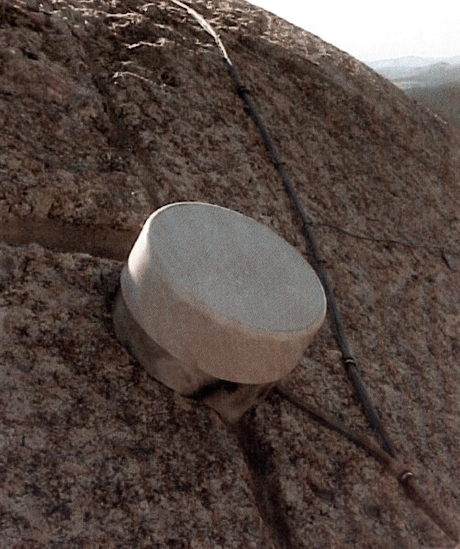 One of many round sensor covers attached to granite on the back of George Washington's sculpted head on Mount Rushmore.  A gray cable runs from the lower right corner of the image to the upper middle where the curve of the top of the head is visible.