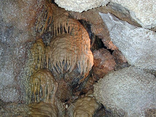 Knobby, light-colored nailhead spar calcite crystals cover a cave wall on the right side of the image. Caramel-colored dripstone formations that look like folded curtains cascade over rocks on the left side of the image in Jewel Cave.