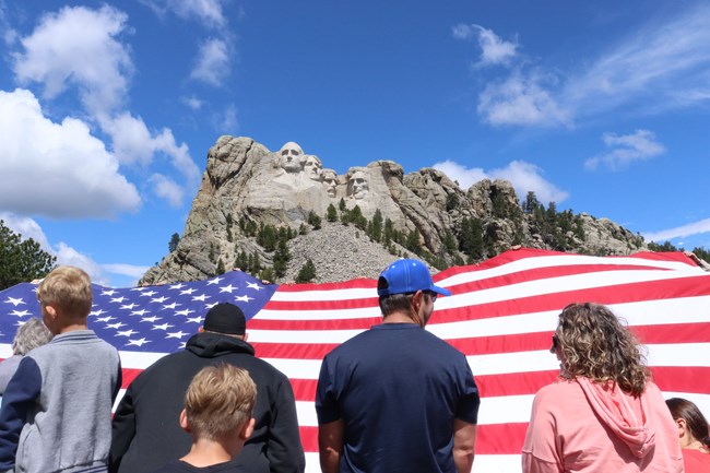 Visitors lift the American flag with the sculpture of Mount Rushmore in the background.