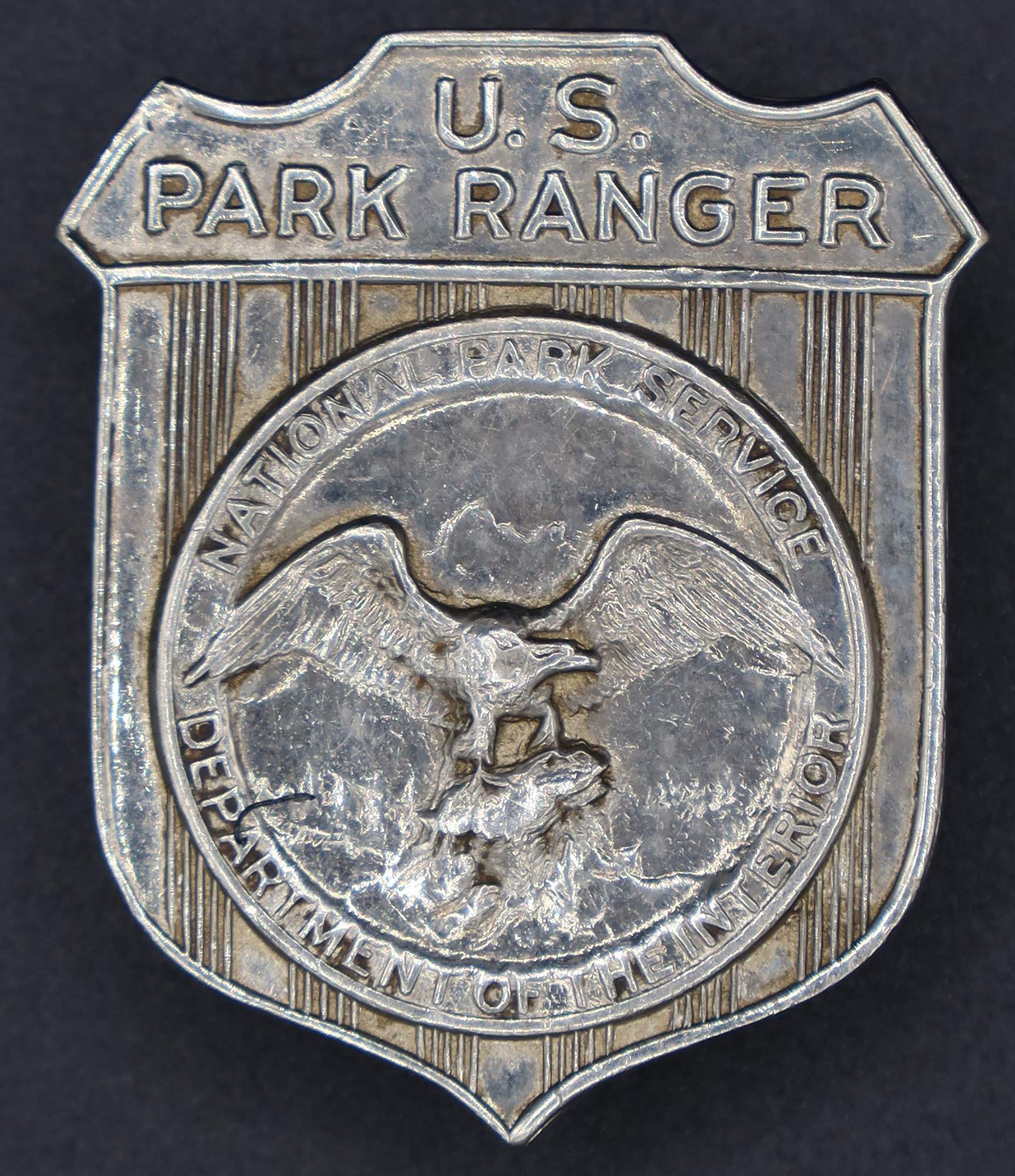 Silver shield-shaped badge marked U.S. Park Ranger. The raised round seal in the middle has an eagle looking to its left.