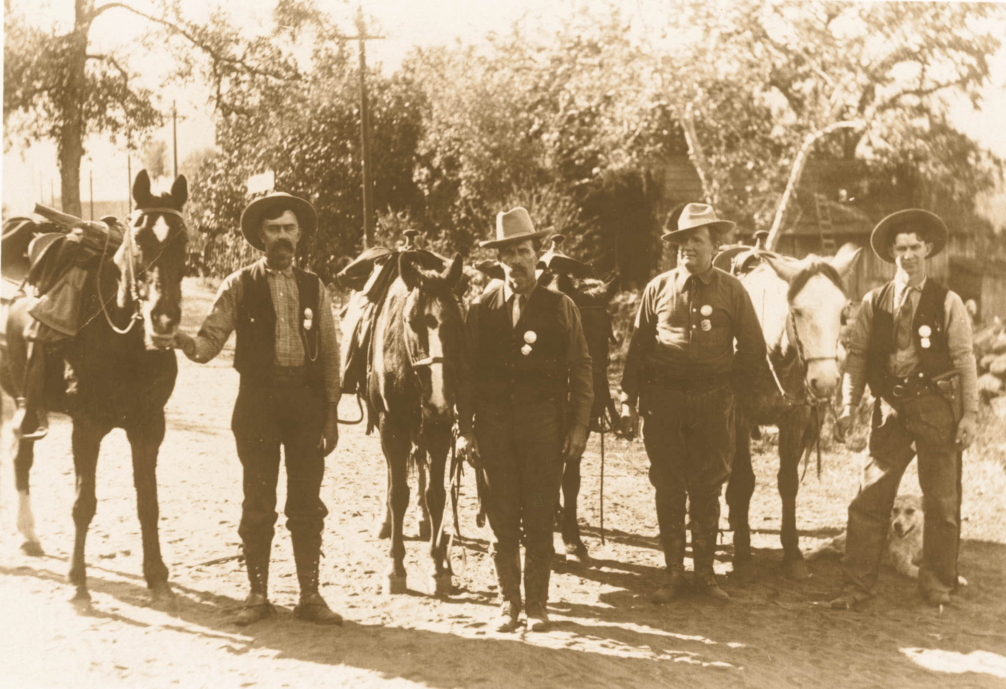 Four men in cowboy-type riding clothes stand next to horses. Each wears a round badge above a shield shaped badge, on a vest or shirt.