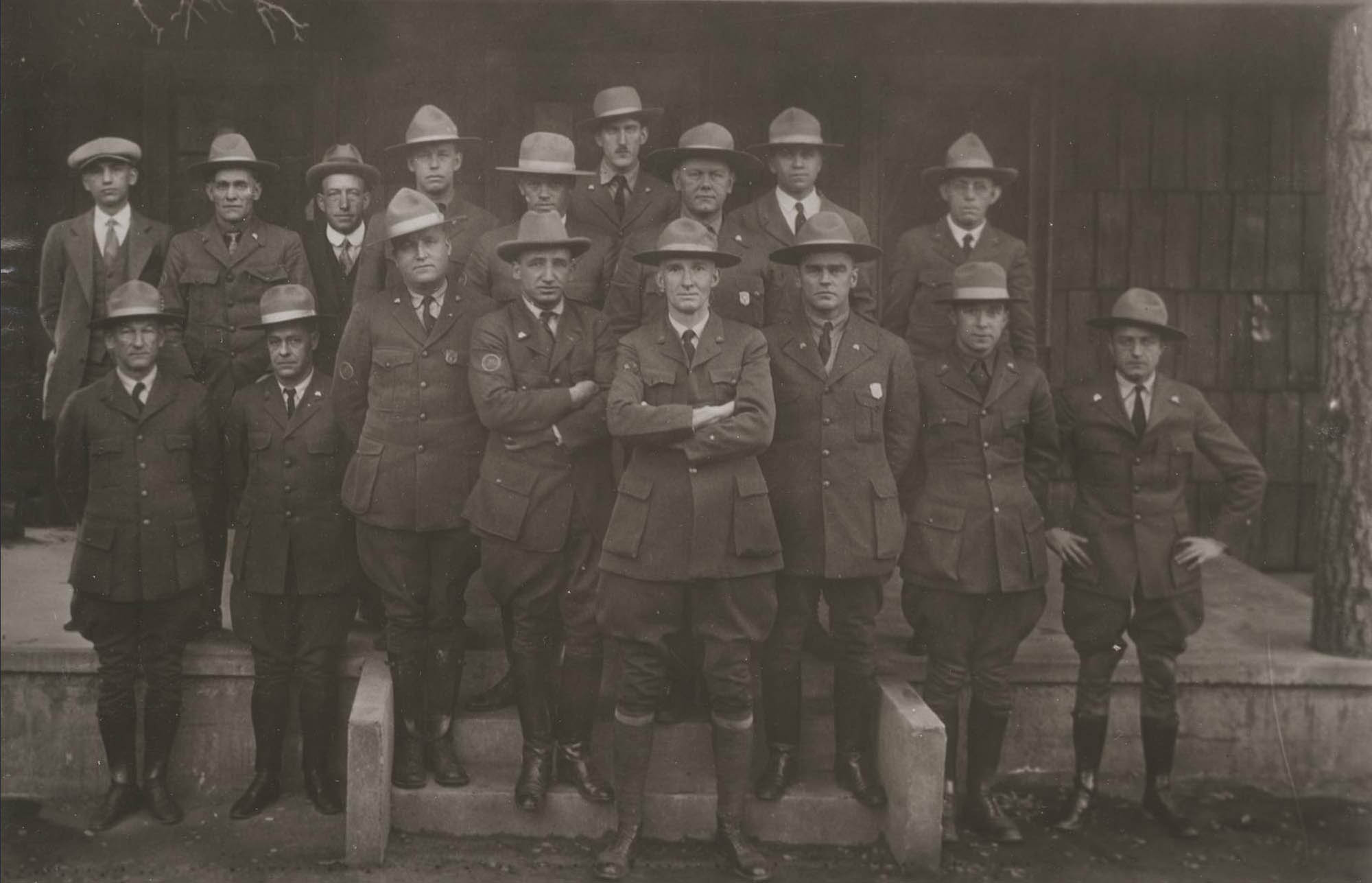 Seventeen men pose in in front of a building, 15 in NPS uniforms with broad brim hats. Shield-shaped badges are worn only by three men.