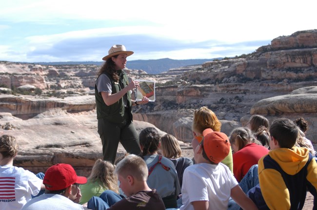 A ranger wearing green clothes holds a photo up to show a group of children. Sandstone rocks and canyons are behind the ranger.
