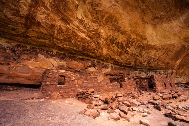 Reddish brown stone structures with piles of rocks in front of them. The sturctures are under a canyon overhang.