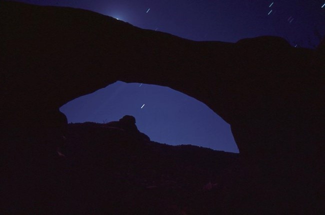 white star trails on a dark blue sky around the silhouette of a natural bridge