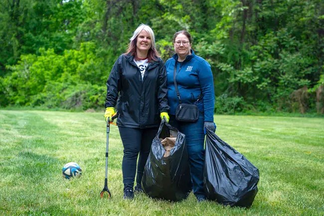 Two volunteers stand with a trash bag and picker on a grassy field.