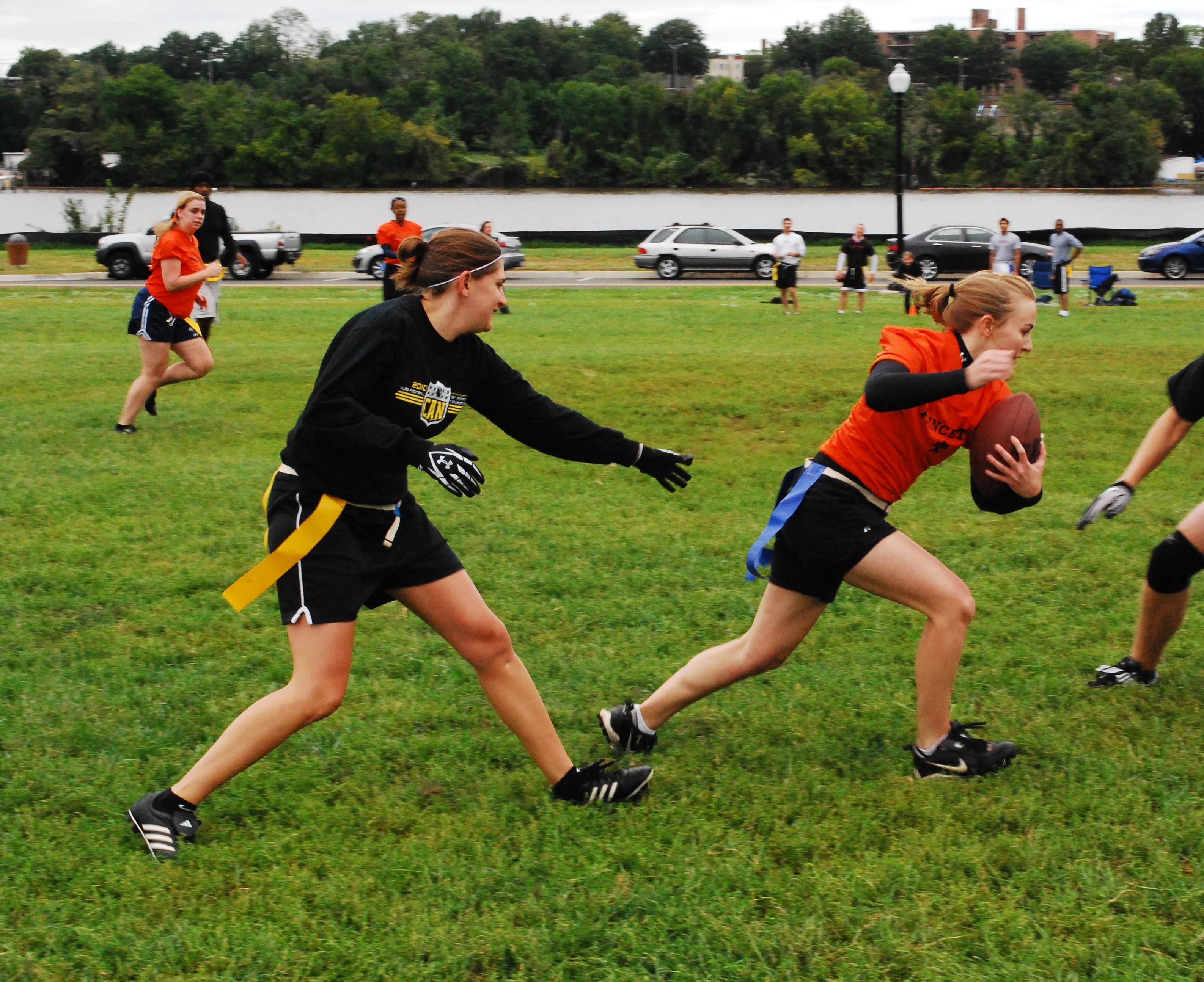 A woman in a red jersey runs with a football while a woman in a black jersey reaches to grab her flag during a flag football game on a grassy field.
