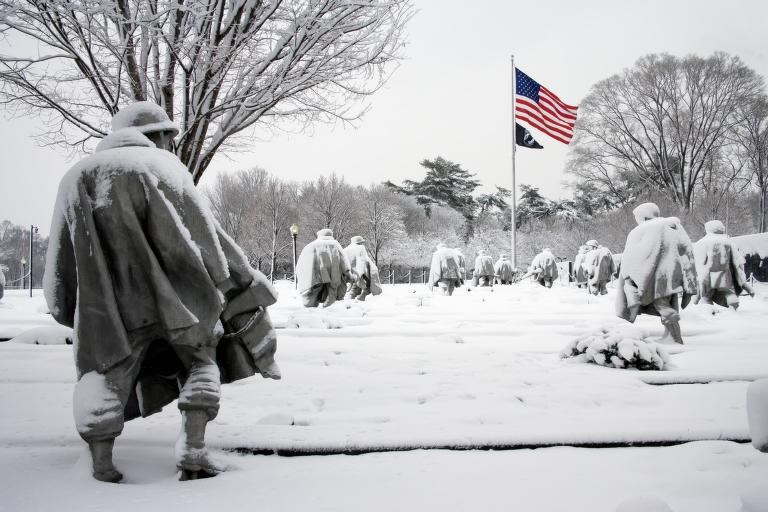 Stainless steel soldiers stand in column with the American Flag flying high at head, all of them under a blanket of snow.