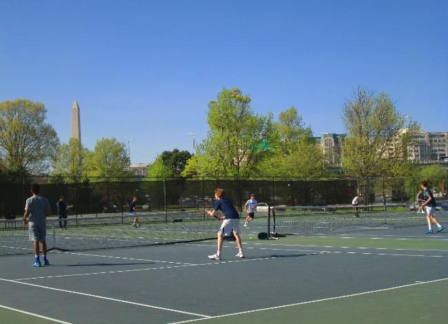Several tennis students playing tennis on an outdoor tennis court. The Washington Monument is in the background.