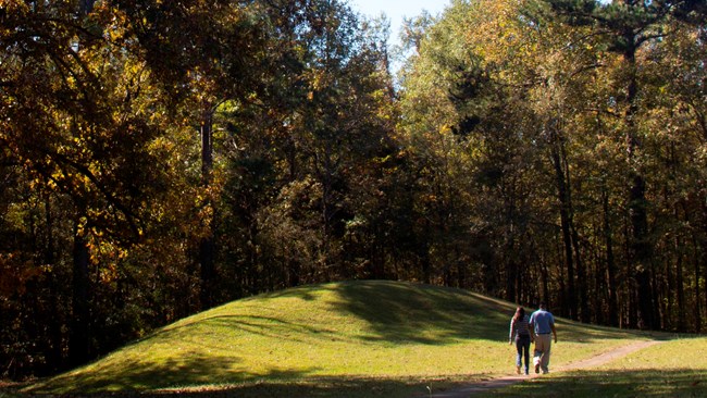 Two people walk on a path next to a grassy hill.