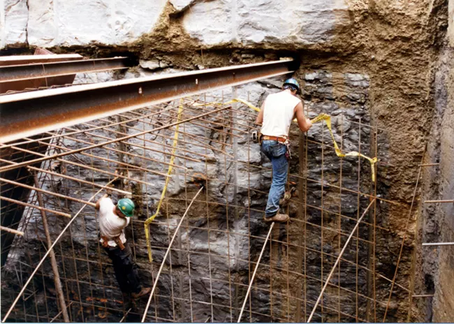 Two construction workers stand inside metal scaffolding on a cliffside.