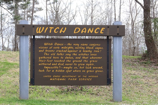 A large brown sign with header board Witch Dance, routed and painted yellow. Below the header is text explaining the site.