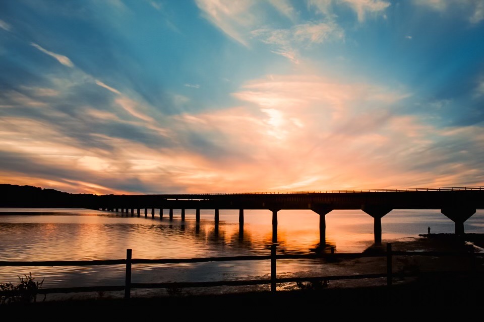The silhouette of a bridge at sunset spanning a wide river. The bridge stands on singular pilar supports. The sky is pink and blue with swirling clouds.