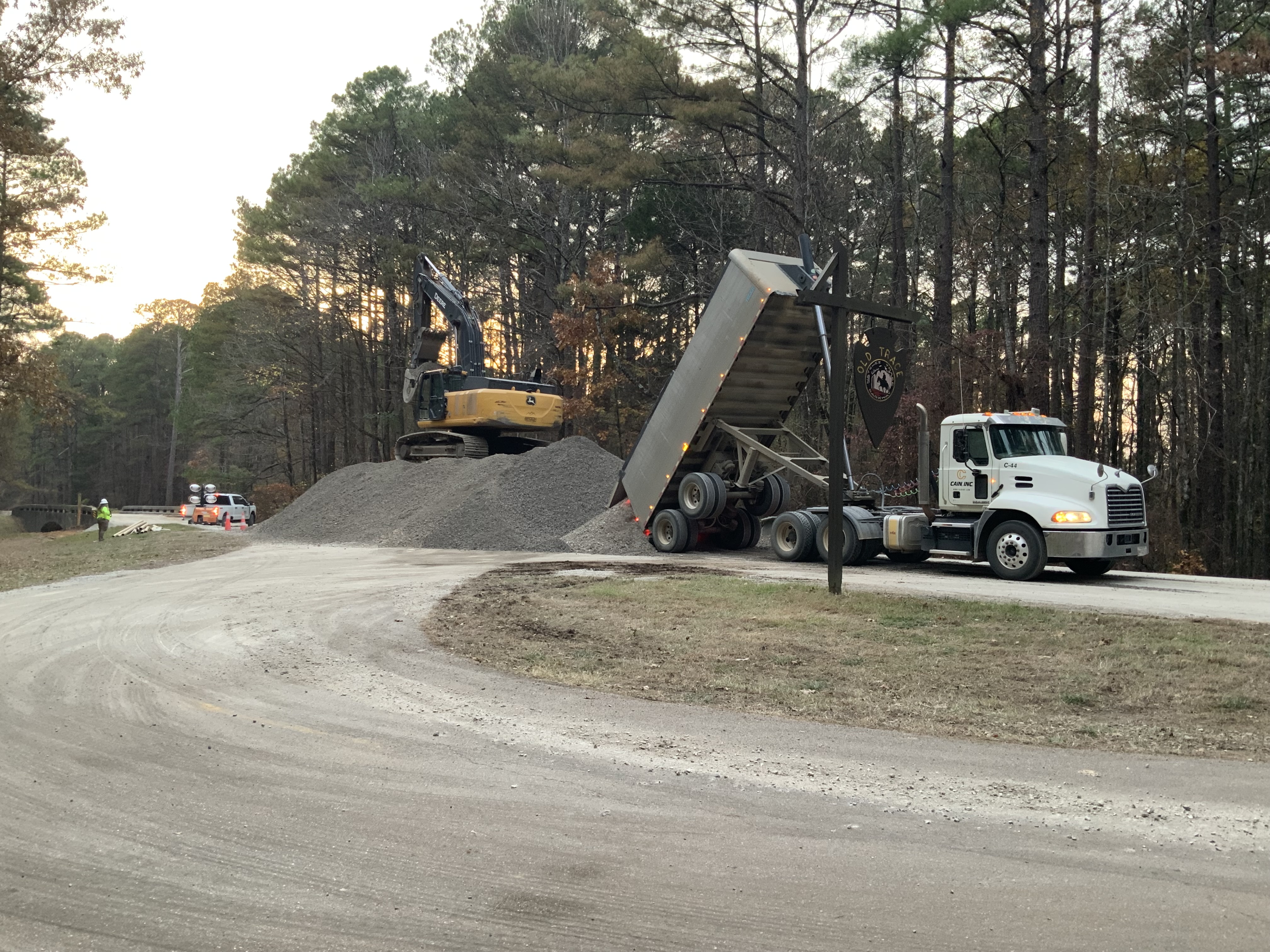 The pull-off site for a section of Old Trace. In the background, there is an excavator atop a large pile of gravel. A dump truck unloads more gravel onto the pile.