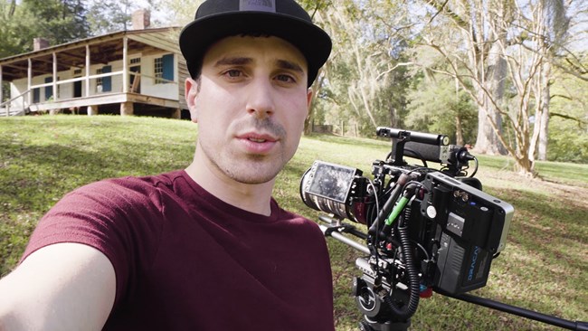 A closeup of a man with a camera with an old cabin in the background.
