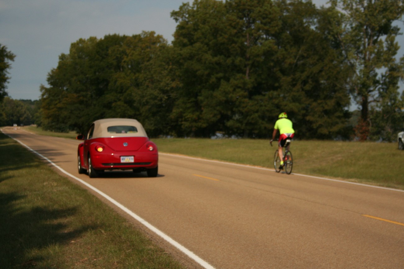 A red car passed a bicyclist with a bright florescent yellow shirt.