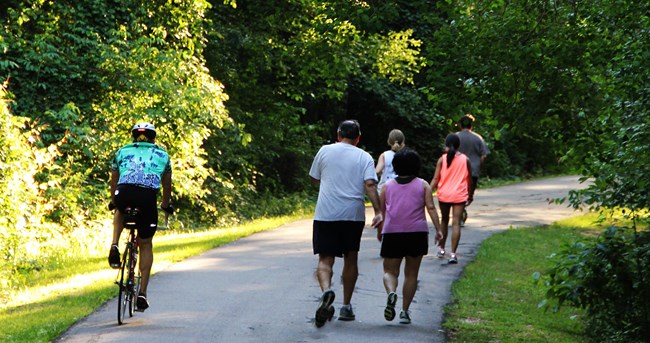 A bicyclist passes pedestrians on a paved path through a wooded area.