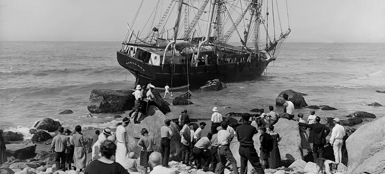 A crowd gathers on a rocky shore, watching the grounded whaling ship Wanderer, which rests on rocks in shallow water. Some men climb the rocks, attempting to assist, while the ocean stretches out in the background.