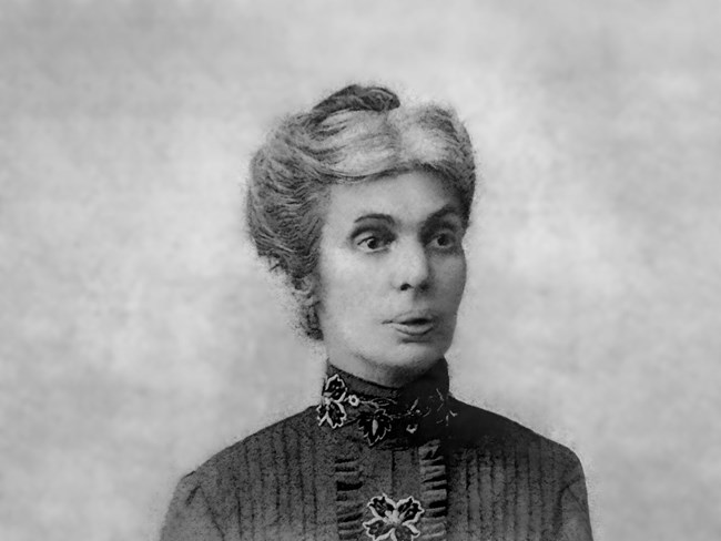 Black-and-white portrait of Maria F. Soares, a woman with light-colored hair styled in an updo, wearing a high-collared, dark patterned dress with floral embroidery.