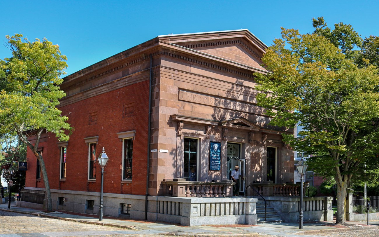 The image shows the New Bedford Whaling National Historical Park Visitor Center, located in the historic Third District Courthouse building. The red brick building has stone detailing with "THIRD DISTRICT COURT". A park ranger stands in front of the door