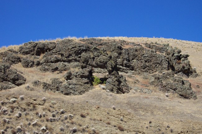 Brown grassy hillside with black rock outcrops and a rock arch in the middle.