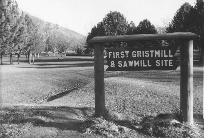 Historic black and white photo of a wooden sign labeled Fist Gristmill and Sawmill Site in front of a grassy field with leafy trees.