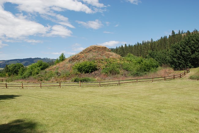 A large mound of rock encircled by a wooden fence and surrounded by green grass.