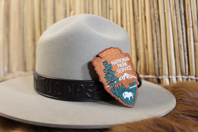 A grey hat with a brown hat band that says US NPS and an NPS arrowhead patch sitting on a brown fur pelt with a tan plant reed in the background.