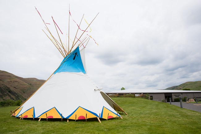 A large blue, yellow, red, and white painted tipi set-up with long straight poles on a grassy field in front of a grey park visitor center