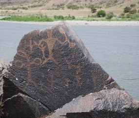 Rock with large petroglyphs of what appear to be stick figures - some are holding items that look somewhat like dumbbells.
