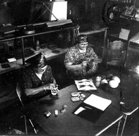 Two women sitting at a table, weaving bags, in an old store.