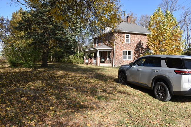 A silver car is parked on a mown lawn in front of a two story brick house.