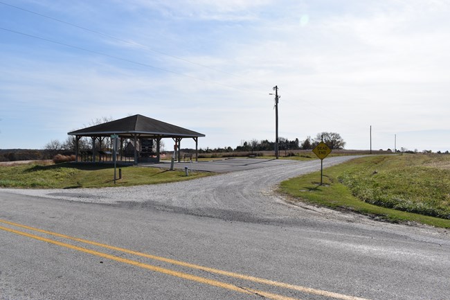 A paved road intersects a gravel entrance road that turns right. It leads to an open-sided pavilion and parking lot.