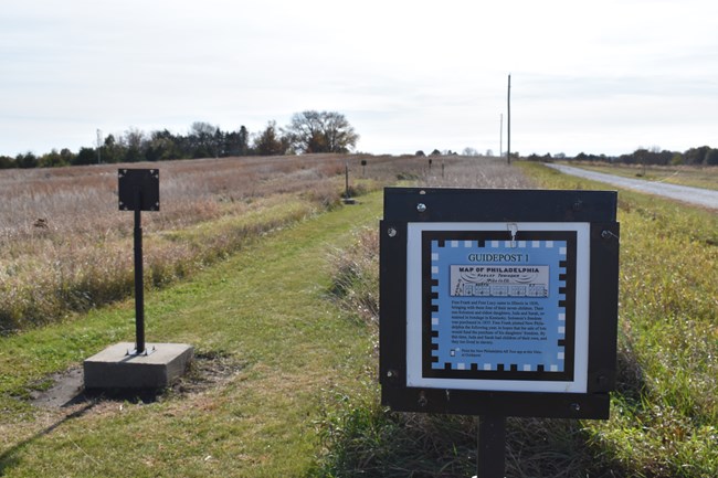 A metal signpost with text information stands at the entrance to a trail through an open field.