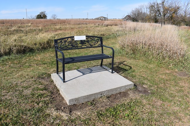 A metal bench on a raised conrete block, in the middle of a field.