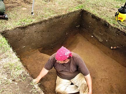 A man wearing dirt covered clothes carefully digging in a brown dirt archaeological excavation pit