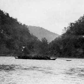 A small shallow flat wooden boat on a wide river between two tree covered hills
