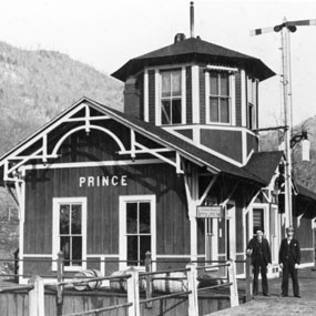 A wooden train depot with the name Prince and two railroad workers standing in front of it