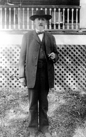 An older man with a white beard wearing an old fashioned suit, bow tie, and hat holding a cigar standing in front of a wooden porch.