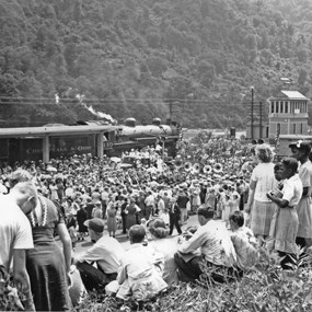 A large crowd of people on a hill outside a train station with a steam locomotive at it. A band playing music is next to the station.