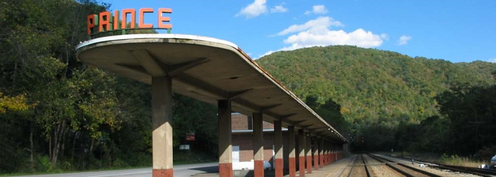 Covered railroad platform with an attached station that says Prince next to railroad tracks with green mountains in the background.