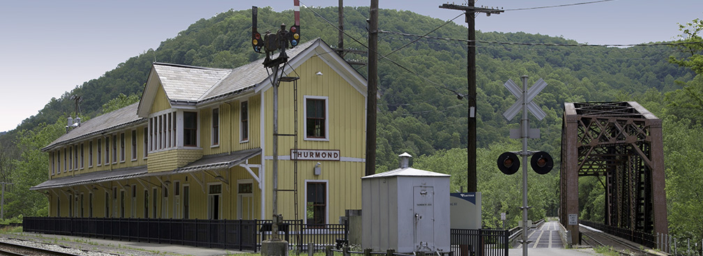A long yellow wooden building next to railroad tracks with a metal railroad bridge next to it
