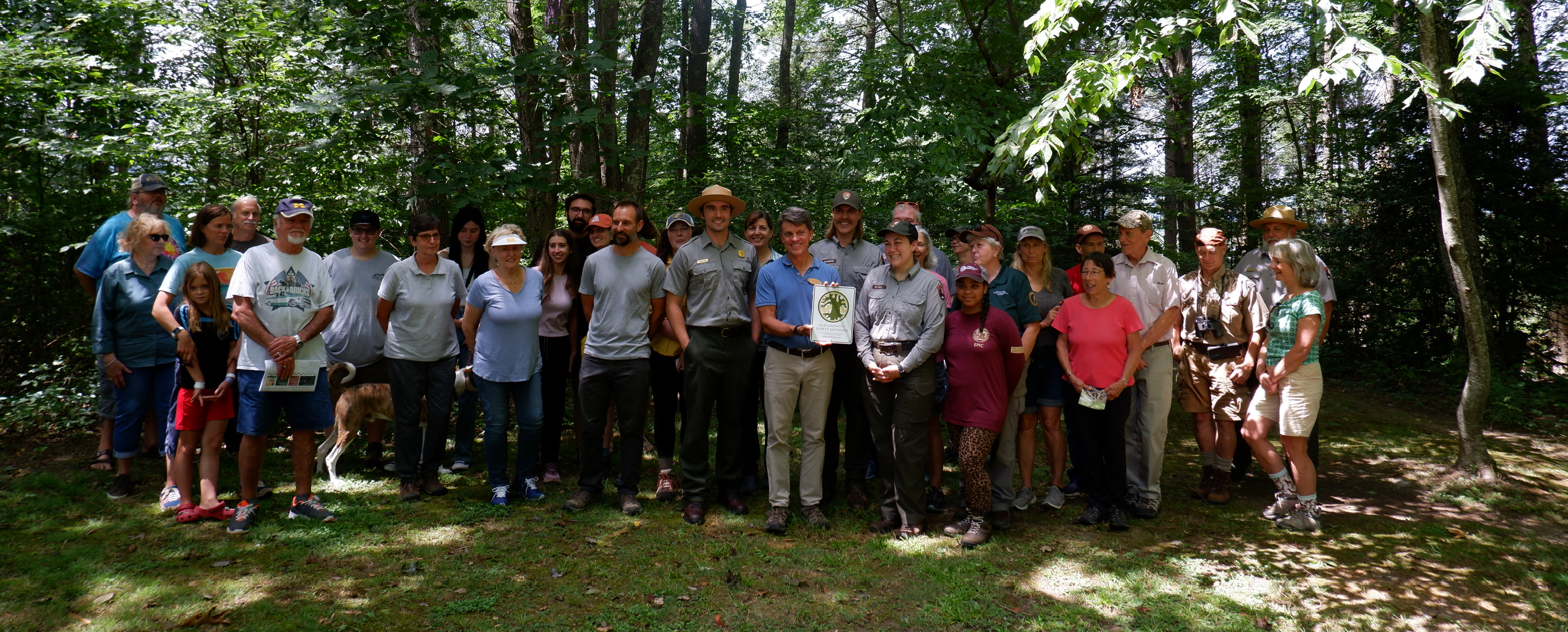 A group of people with 3 park rangers posing in front of trees in a forest. In the middle of the group are 3 park rangers and a person holding a white and green certificate